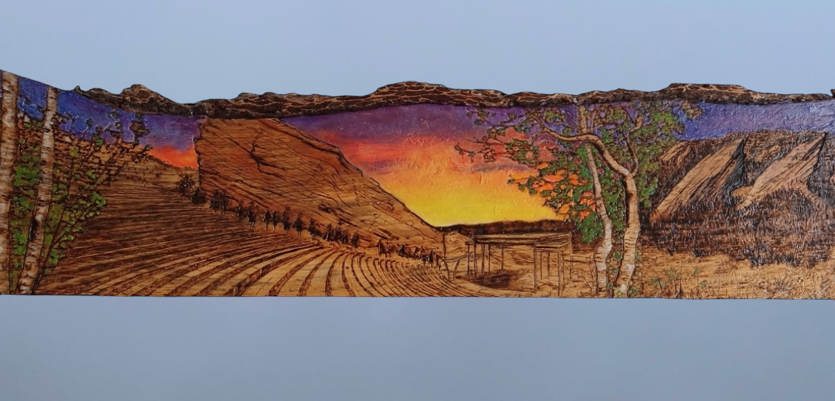 Wood-burned and painted artwork on a reclaimed wood slat with bark backing, depicting Colorado’s Red Rocks Amphitheatre and Boulder Flatirons. The landscape features a vibrant sunset sky in orange, yellow, purple, and blue. Combines pyrography and painting techniques to create a textured, nature-inspired tribute to iconic Colorado landmarks.