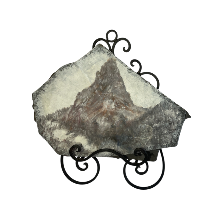 A hand-painted landscape on a piece of natural slate depicts a granite peak rising out of the fog above the pines below. The painting blends earthy greens, blues, and grays, capturing the serenity of the scene. The slate is mounted on an ornate black metal stand with scrollwork details, adding elegance and contrast.